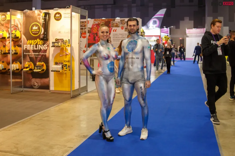 Two models covered in futuristic body paint walk the exhibition floor at EroExpo Moscow, where creativity and sensuality blend in unique artistic expressions.