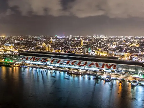 Nachtzicht op Amsterdam met verlichte gebouwen en het Centraal Station aan het water, met grote letters “AMSTERDAM” op het dak.
