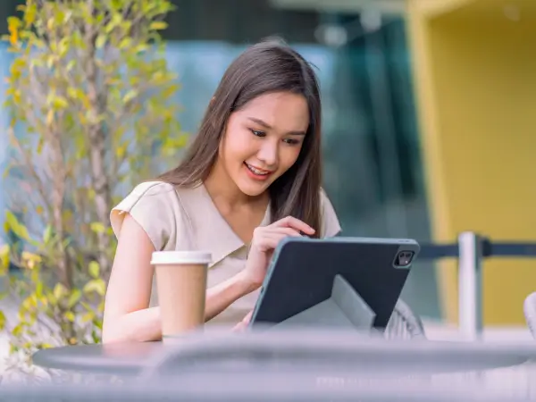 หญิงสาวชาวไทยนั่งอยู่ที่โต๊ะกลางแจ้ง ยิ้มขณะใช้แท็บเล็ต สื่อถึงการสื่อสารธุรกิจออนไลน์และความเป็นมืออาชีพของ Gaia Sex Tech
