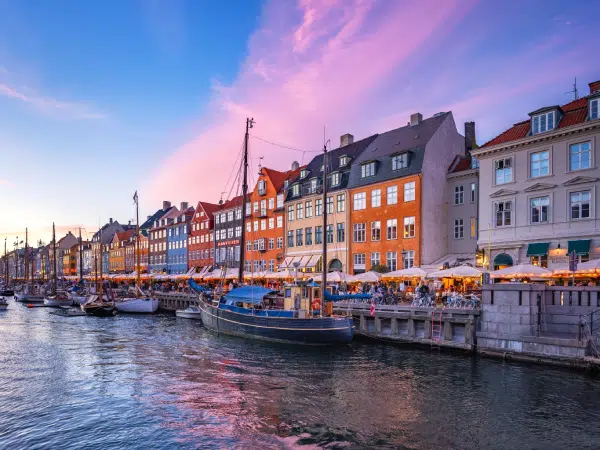 Farverige bygninger og både langs Nyhavn i København ved solnedgang, med mennesker der nyder aftenen ved vandet.