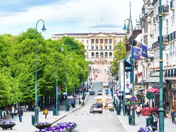 Utsikt opp Karl Johans gate mot Det kongelige slott i Oslo en vårdag, med gående og butikker langs gaten.