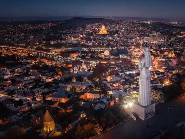 Aerial night view of Tbilisi with the Mother of Georgia statue overlooking the illuminated city skyline.