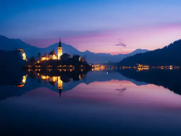 Večerni pogled na Blejsko jezero z osvetljenim otokom in cerkvijo, odsev v mirni vodi – Slovenija, uvodna slika strani »O nas« podjetja Gaia Sex Tech.