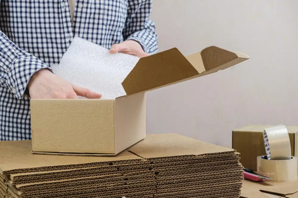 Person packing a plain corrugated shipping box with protective foam—neutral outer carton for fulfillment-ready delivery.