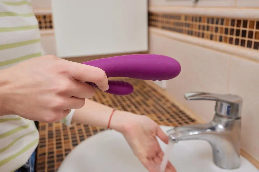 A person cleaning a waterproof vibrator under running water in a sink, demonstrating the proper cleaning method for waterproof sex toys.