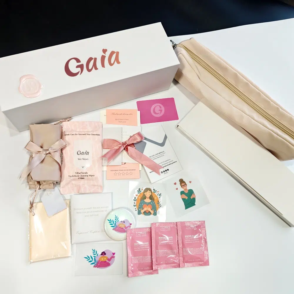 Gaia adult toys packaging flat lay with branded white gift box, zip pouch, manuals and cards, ribbon bundle, wipes, stickers, accessory boxes, and water-based lubricant sachets.