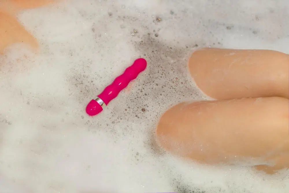 Pink waterproof vibrator floating in a bubble bath beside a person’s legs, illustrating bath-use water play.