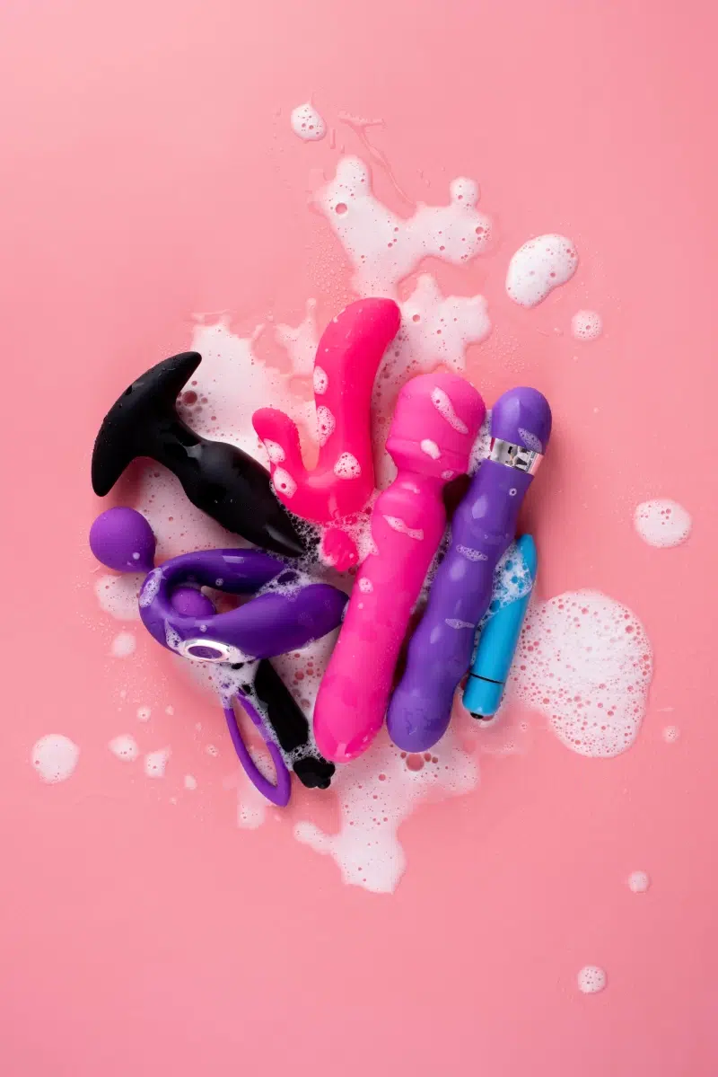 A variety of waterproof sex toys in pink, purple, and black, surrounded by bubbles on a pink background, showcasing their ability to handle water exposure.
