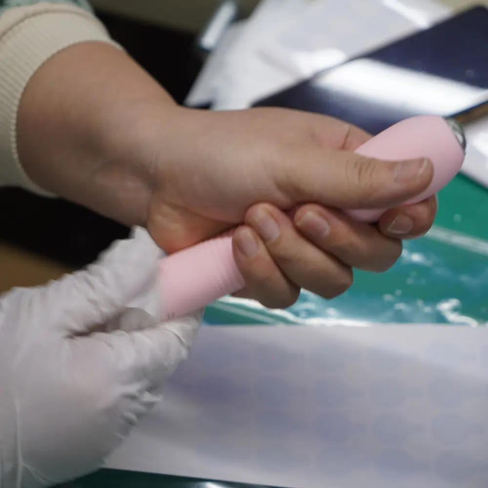 Gloved hands inspecting a pink vibrator during final testing and labeling.