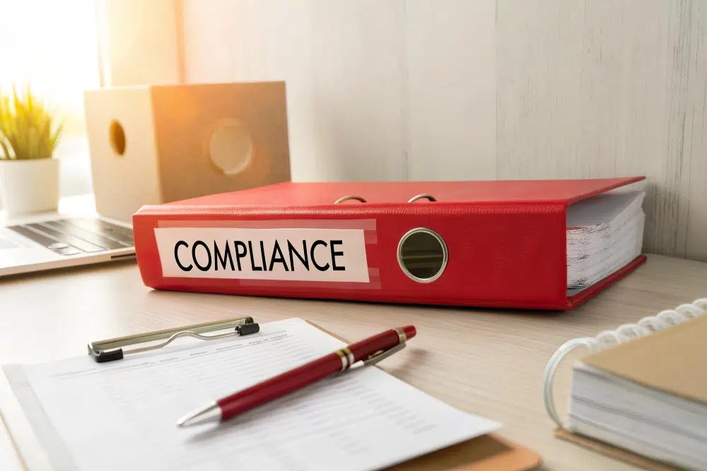 Red binder labeled “COMPLIANCE” on a desk with paperwork and a pen, representing compliance documents and standards.